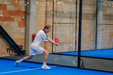 Riga, Letonya - 24 Ocak 2026: Padel turnuvası. Beyaz elbiseli bir erkek sporcu, kapalı bir alanda mavi bir padel kortunda raketle topa vuruyor.