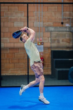 Riga, Letonya - 24 Ocak 2026: Padel turnuvası. Açık tişörtlü ve şortlu bir erkek sporcu mavi bir iç sahada ağ ile bir raket sallıyor.