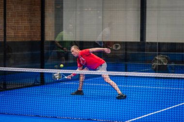 Riga, Letonya - 24 Ocak 2026: Padel turnuvası. Kırmızı tişörtlü bir erkek atlet arka planda ağ ile mavi bir padel kortta ters vuruş yapıyor.