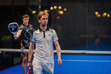 Riga, Letonya - 24 Ocak 2026: Padel turnuvası. Mavi kortta iki erkek oyuncu, biri beyaz Nike tişörtlü, diğeri siyah tişörtlü.