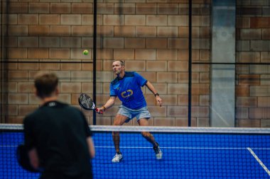 Riga, Letonya - 24 Ocak 2026: Padel turnuvası. Mavi tişörtlü ve şortlu erkek padel oyuncusu mavi bir kortta kapalı alanda oynanan maçta topu bekliyor.