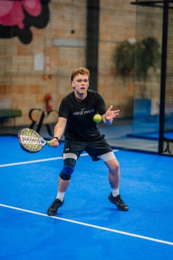 Riga, Letonya - 24 Ocak 2026: Padel turnuvası. Nike Pro 'daki genç erkek sporcu, mavi kapalı bir padel kortunda camdan duvarları olan bir top bekliyor.