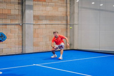 Riga, Letonya - 24 Ocak 2026: Padel turnuvası. Parlak turuncu tişörtlü erkek padel oyuncusu, topu camdan duvarları olan mavi bir kortta geri verecek pozisyonda.
