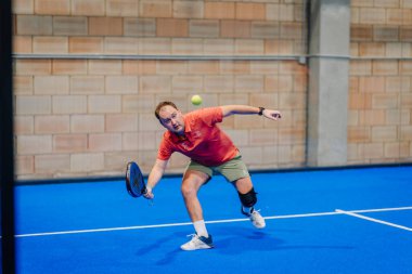 Riga, Letonya - 24 Ocak 2026: Padel turnuvası. Turuncu tişört giyen erkek padel oyuncu mavi kortta dizlik ile topa vurmak için hamle yapıyor.