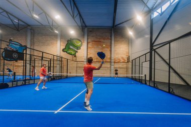 Riga, Letonya - 24 Ocak 2026: Padel turnuvası. Renkli duvar resimleri olan iyi ışıklandırılmış bir tesisin içindeki mavi bir kortta padel oynayan iki kişi.