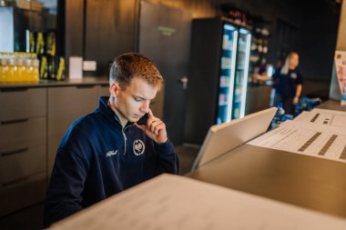 Riga, Letonya - 24 Ocak 2026: Padel turnuvası. Donanma polo tişörtlü genç resepsiyonist, padel spor tesisinde bir resepsiyonda dizüstü bilgisayar kullanıyor.