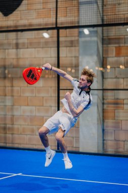 Riga, Letonya - 24 Ocak 2026: Padel turnuvası. Beyaz elbiseli bir erkek atlet, kırmızı bir padel raketle mavi bir kortta topa vuruyor.