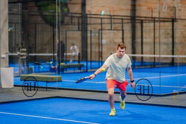 Riga, Letonya - 24 Ocak 2026: Padel turnuvası. Açık tişörtlü ve şortlu bir erkek atlet, mavi bir kortta cam bir duvar ve görünür yansımalarla padel oynar