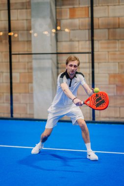 Riga, Letonya - 24 Ocak 2026: Padel turnuvası. Beyaz kıyafetli erkek sporcu, mavi bir kortta kırmızı bir padel raketle topa vurabilir.