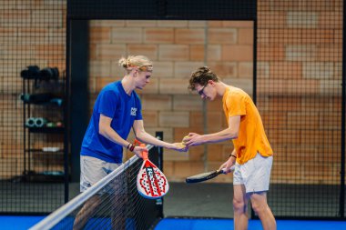 Riga, Letonya - 24 Ocak 2026: Padel turnuvası. Mavi ve turuncu tişörtlü iki erkek oyuncu kapalı bir sahada mavi döşemeli topları değiş tokuş ediyor.