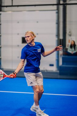 Riga, Letonya - 24 Ocak 2026: Padel turnuvası. Mavi tişörtlü ve şortlu erkek padel oyuncu mavi sahada seyircilerle topa vuracak pozisyonda.