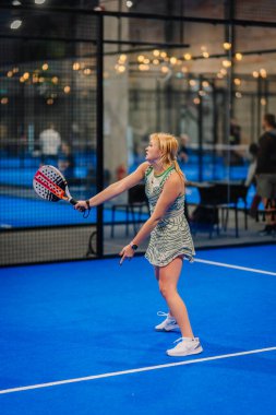 Riga, Letonya - 24 Ocak 2026: Padel turnuvası. Desenli elbiseli bayan padel oyuncu, mavi sahada seyircilerin gözü önünde topa vurmaya hazırlanıyor.