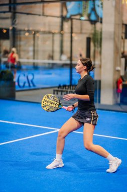 Riga, Letonya - 24 Ocak 2026: Padel turnuvası. Siyah bluzlu ve şortlu kadın padel oyuncu canlı mavi kortta camdan duvarlarla topa vurmaya hazırlanıyor.