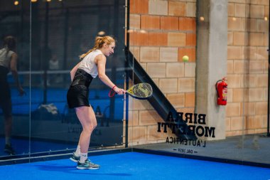 Riga, Letonya - 24 Ocak 2026: Padel turnuvası. Beyaz atlet ve siyah şortlu bayan sporcu kapalı bir padel kortunda raketle topa vurmaya hazırlanıyor.