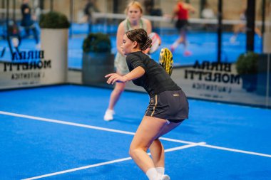 Riga, Letonya - 24 Ocak 2026: Padel turnuvası. Siyah elbiseli bayan padel atlet topa canlı bir mavi sahada cam panellerle vuracak.