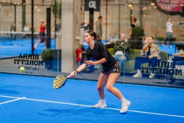 Riga, Letonya - 24 Ocak 2026: Padel turnuvası. Siyahlı bayan atlet, canlı bir mavi sahada seyircilerle birlikte padel topuna vurmaya hazırlanıyor.