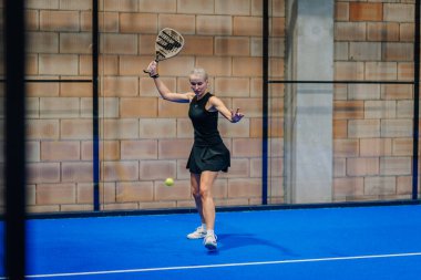 Riga, Letonya - 24 Ocak 2026: Padel turnuvası. Siyah elbiseli kadın padel oyuncu mavi kortta duvara vurmak için raket sallıyor.