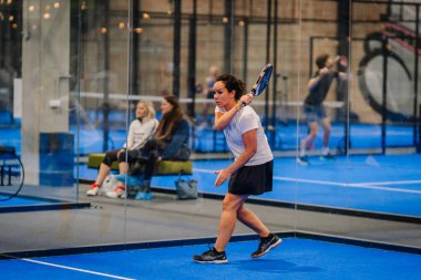 Riga, Letonya - 24 Ocak 2026: Padel turnuvası. Beyaz tişörtlü ve siyah etekli bayan padel oyuncu, mavi sahada seyircilerle topa vurmaya hazırlanıyor.