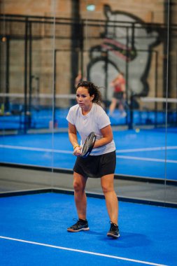 Riga, Letonya - 24 Ocak 2026: Padel turnuvası. Beyaz tişörtlü ve siyah şortlu bir bayan atlet mavi kortta bir padel topuna vurmaya hazırlanıyor.