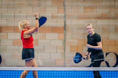 Riga, Letonya - 24 Ocak 2026: Padel turnuvası. İki bayan oyuncu kapalı alanda maç yapıyor, biri kırmızı bluzlu kutlama yaparken diğeri oyuna odaklanıyor.