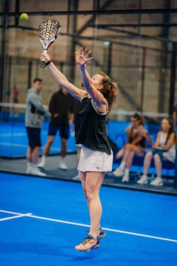 Riga, Letonya - 24 Ocak 2026: Padel turnuvası. Siyah askılı ve beyaz şortlu bayan padel oyuncu mavi sahada topa vurmaya hazırlanıyor.