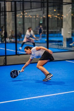 Riga, Letonya - 24 Ocak 2026: Padel turnuvası. Gri gömlekli ve siyah etekli bayan sporcu, mavi bir padel kortunda kürekle topa vurmak için eğiliyor.