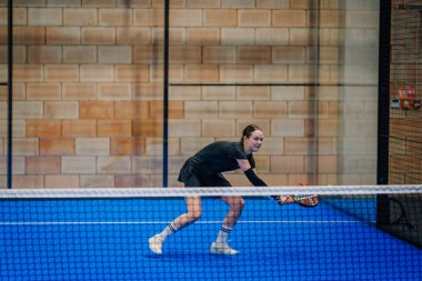 Riga, Letonya - 24 Ocak 2026: Padel turnuvası. Siyah kıyafetli kadın padel oyuncu mavi sahada topa vurmak için çömeliyor.