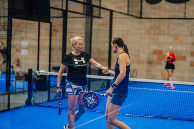 Riga, Letonya - 24 Ocak 2026: Padel turnuvası. Padel maçında iki bayan oyuncu, mavi sahada bir top paylaşıyor. Ağ ve duvarlar görünüyor.