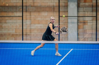 Riga, Letonya - 24 Ocak 2026: Padel turnuvası. Siyah elbiseli kadın padel oyuncu mavi kortta camlı duvarlı raketiyle topa vurmaya hazırlanıyor.