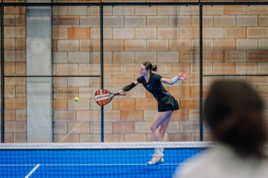 Riga, Letonya - 24 Ocak 2026: Padel turnuvası. Siyah kıyafetli kadın padel oyuncu kapalı bir spor tesisinde mavi kortta raketle topa vuruyor.