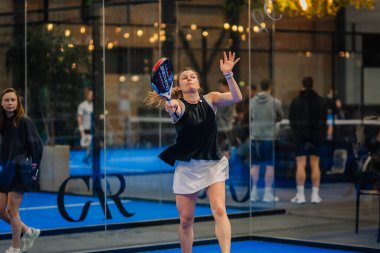 Riga, Letonya - 24 Ocak 2026: Padel turnuvası. Siyah bluzlu ve beyaz etekli kadın padel oyuncu topu camdan duvarları olan kapalı bir sahada servis ediyor.