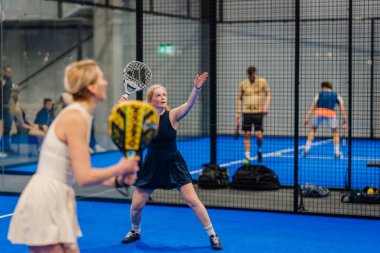 Riga, Letonya - 24 Ocak 2026: Padel turnuvası. İki kadın mavi kortta padel oynuyor, biri beyaz elbiseli, diğeri koyu renk elbiseli, ağlı.