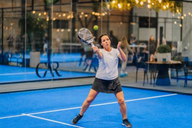 Riga, Letonya - 24 Ocak 2026: Padel turnuvası. Beyaz tişörtlü ve siyah etekli bayan padel oyuncu mavi bir kortta yansımalarla topa vurabilir.