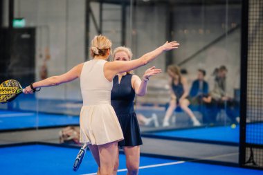 Riga, Letonya - 24 Ocak 2026: Padel turnuvası. Spor elbiseli iki kadın, arka planda cam duvarlar ve izleyiciler olan mavi bir padel kortta sarılıyorlar.