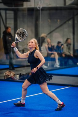 Riga, Letonya - 24 Ocak 2026: Padel turnuvası. Koyu renk elbiseli bayan padel atlet arka planda izleyiciler izlerken mavi kortta raket sallıyor.