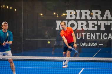 Riga, Letonya - 24 Ocak 2026: Padel turnuvası. İki erkek Padel oyuncusu mavi kortta bir maç yapıyor, biri turuncu tişört giyiyor, diğeri mavi.