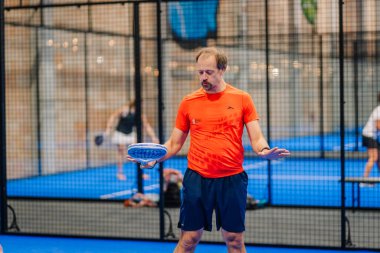 Riga, Letonya - 24 Ocak 2026: Padel turnuvası. Turuncu tişörtlü erkek padel oyuncusu mavi bir sahada mavi raketi ağla tutarken tekniğini gösteriyor.