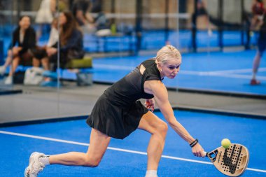 Riga, Letonya - 24 Ocak 2026: Padel turnuvası. Siyah elbiseli bayan padel oyuncu, arka planda seyircilerin olduğu mavi bir sahada topu iade etmek için geriniyor.