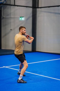 Riga, Letonya - 24 Ocak 2026: Padel turnuvası. Bej tişörtlü ve siyah şortlu bir erkek sporcu mavi bir sahada topa vurmaya hazırlanıyor.