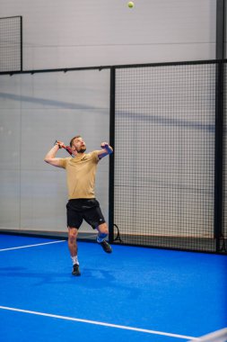 Riga, Letonya - 24 Ocak 2026: Padel turnuvası. Bej tişört giyen erkek padel oyuncu, bir spor tesisi içinde ağlı mavi bir sahada top servis ediyor.