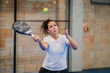 Riga, Letonya - 24 Ocak 2026: Padel turnuvası. Beyaz tişörtlü ve siyah şortlu bir bayan atlet kapalı sahada bir padel topuna vurmaya hazırlanıyor.