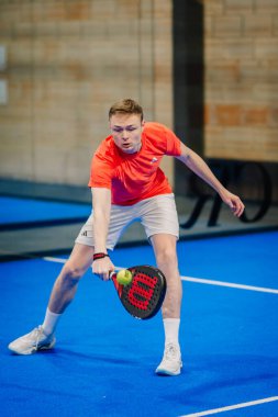 Riga, Letonya - 24 Ocak 2026: Padel turnuvası. Kırmızı tişörtlü ve beyaz şortlu genç padel oyuncusu mavi sahada topa vurmaya hazırlanıyor.