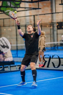 Riga, Letonya - 24 Ocak 2026: Padel turnuvası. Siyah tişörtlü ve şortlu erkek padel oyuncu, mavi sahada izleyicilerle topa vurmaya hazırlanıyor.