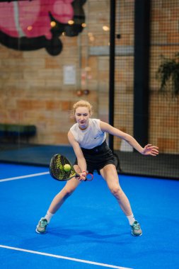 Riga, Letonya - 24 Ocak 2026: Padel turnuvası. Kolsuz tişörtlü ve şortlu bayan padel atlet mavi iç sahada topa vuracak pozisyonda.