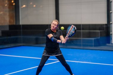 Riga, Letonya - 24 Ocak 2026: Padel turnuvası. Siyah elbiseli kadın padel oyuncu mavi bir kortta renkli bir raket sallıyor.