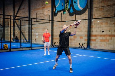 Riga, Letonya - 24 Ocak 2026: Padel turnuvası. Siyah tişörtlü erkek padel oyuncu mavi sahada kırmızı tişörtlü başka bir oyuncuyla topa vurmak için raket sallıyor.