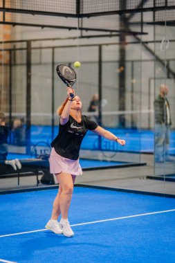 Riga, Letonya - 24 Ocak 2026: Padel turnuvası. Siyah bluzlu ve pembe etekli bayan padel oyuncu mavi bir kortta camdan duvarları ve seyircileri olan bir top sunuyor.