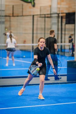 Riga, Letonya - 24 Ocak 2026: Padel turnuvası. Siyah elbiseli bayan sporcu mavi bir padel kortunda topa vurmaya hazırlanıyor.