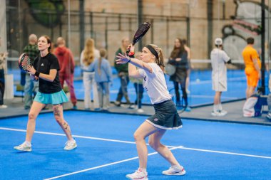 Riga, Letonya - 24 Ocak 2026: Padel turnuvası. Mavi kortta iki bayan padel oyuncusu, biri beyaz gömlekli ve etekli topa vurmaya hazırlanıyor.