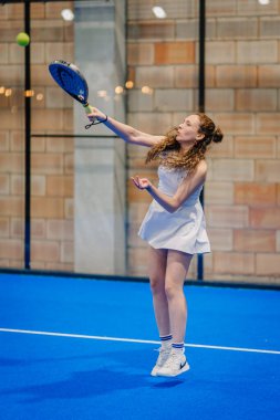 Riga, Letonya - 24 Ocak 2026: Padel turnuvası. Beyaz elbiseli bayan padel oyuncu, mavi kortta cam duvarlı bir baloya hazırlanıyor.